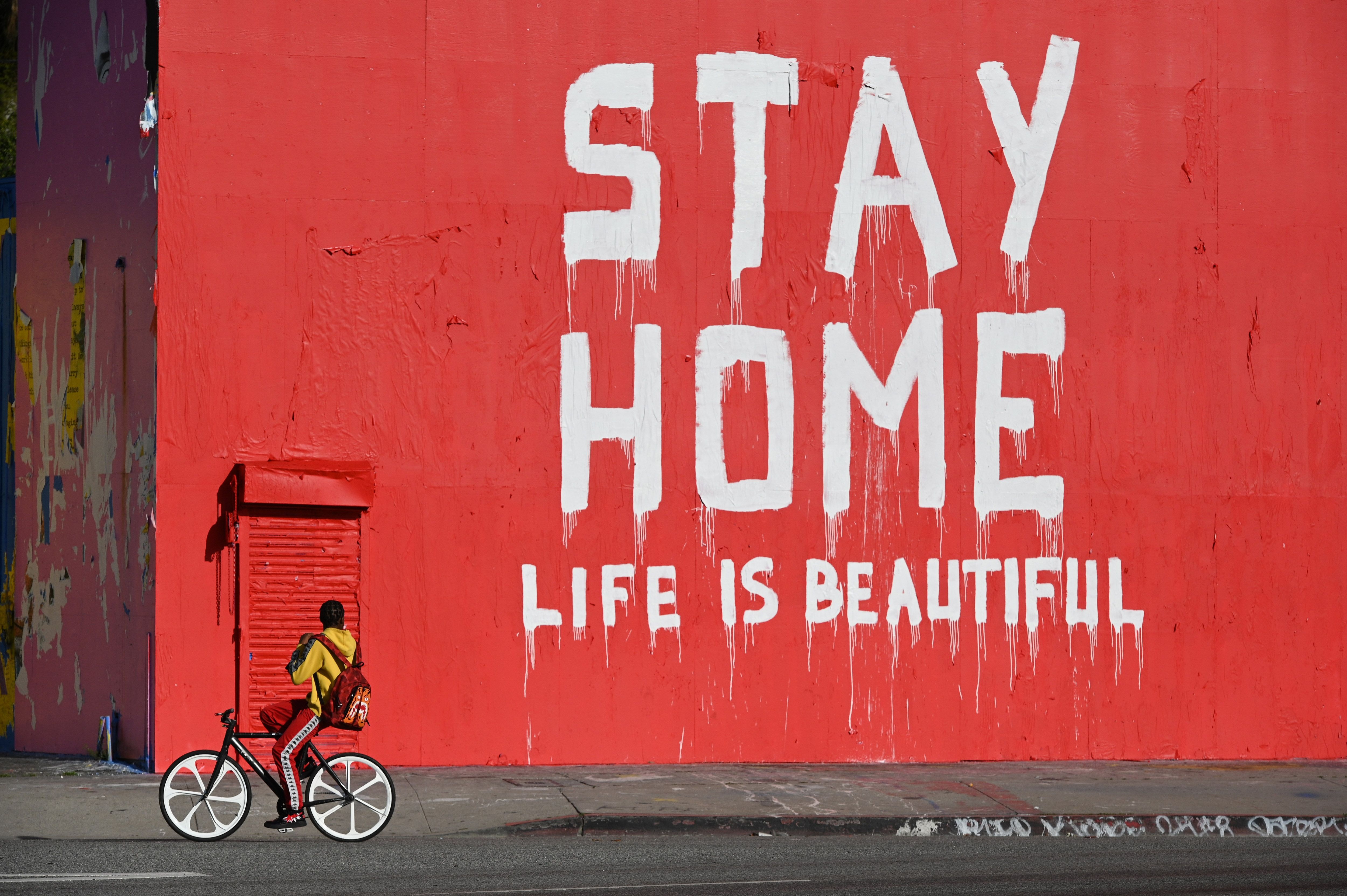 Un hombre pasa en su bicicleta junto a un mural que dice "Quédate en casa / La vida es bella" durante la pandemia de coronavirus (Covid-19) el 3 de abril de 2020 en Los Ángeles, California. (Foto de Robyn Beck / AFP) (Foto de ROBYN BECK / AFP a través de Getty Images)