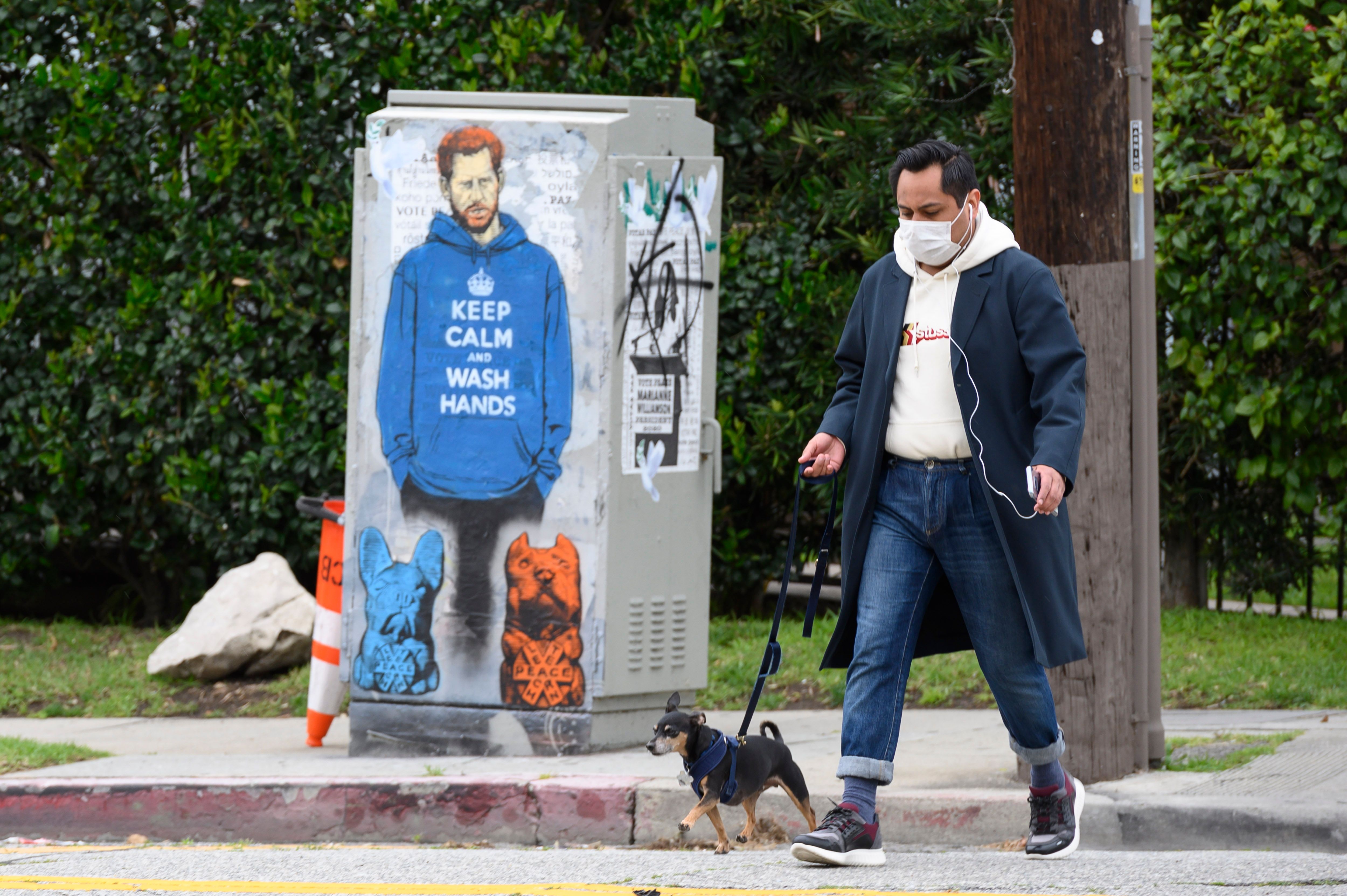 La gente pasa frente al graffiti del Príncipe Harry con una sudadera con capucha que dice "Mantenga la calma y lávese las manos", el 7 de abril de 2020, durante la pandemia de coronavirus en Los Ángeles, California. (Foto de Robyn Beck / AFP) (Foto de ROBYN BECK / AFP a través de Getty Images)