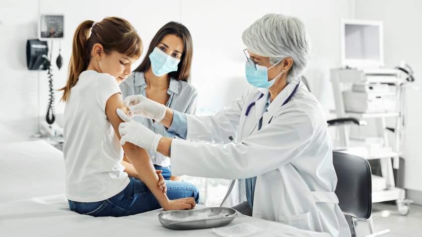 Mature doctor applying bandage on arm of girl. Female pediatrician is examining child sitting on examination table. Family is visiting clinic during COVID-19 outbreak.