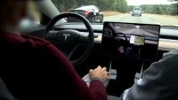 a person sits in the driver seat of a Tesla vehicle, The steering wheel, center monitor and part of the windshield are visible.