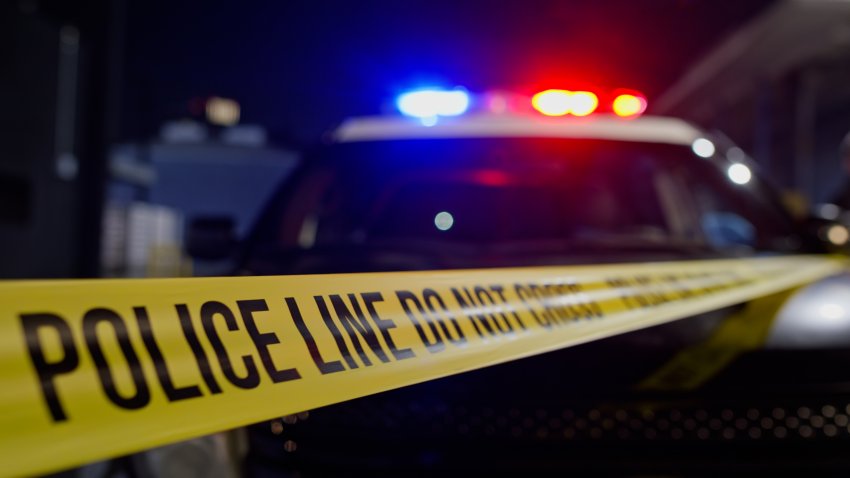 A police crime scene at night with yellow police tape in the foreground reading ‘POLICE LINE DO NOT CROSS’ and emergency vehicles with flashing red and blue lights in the background. The scene is illuminated by the colorful emergency lighting creating a dramatic atmosphere typical of law enforcement investigations and crime scene documentation.